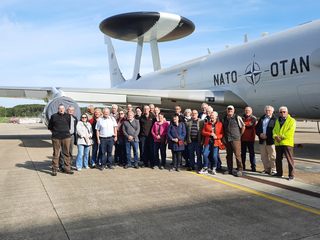 Gruppe von Besuchern posiert vor einem großen weißen NATO AWACS Überwachungsflugzeug auf einem Flugfeld.