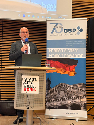 Ein Mann spricht ins Mikrofon an einem Rednerpult mit 'Stadt Bonn' Schild, neben einem GSP-Banner mit Flagge und Reichstag.
