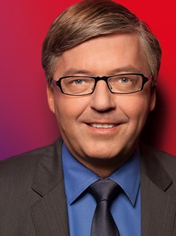 Kopfportrait eines lächelnden Mannes mittleren Alters mit Brille, blauem Hemd, Anzug und Krawatte vor rotem und violettem Hintergrund.