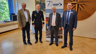 Vier Männer, darunter einer in Marineuniform, posieren in einem Raum mit GSP-Banner und einer großen Kompassrose an der Wand.