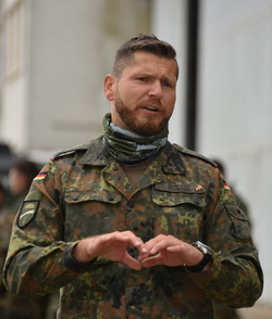 Ein bärtiger Mann in deutscher Tarnuniform und Halstuch formt mit seinen Händen ein Herz und spricht dabei.