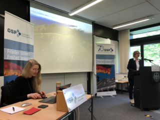 Vortrag in einem Konferenzraum: Eine Frau spricht am Pult, während eine andere am Laptop arbeitet. GSP-Banner und Projektion sind sichtbar.