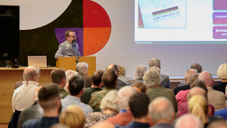 Mann in deutscher Militäruniform spricht an Rednerpult vor Publikum; 'Grundgesetz' auf einer Leinwand zu sehen.