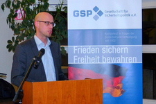 Ein Mann spricht am Pult mit Mikrofon; dahinter ein GSP-Banner mit 'Frieden sichern, Freiheit bewahren' und deutscher Flagge.