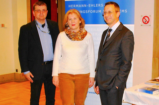 Drei Personen, zwei Männer und eine Frau, stehen nebeneinander vor einem blauen Banner und lächeln in die Kamera.