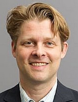 Lächelnder Mann mit hellbraunem Haar, der einen dunklen Anzug und ein gestreiftes Hemd trägt, vor grauem Hintergrund.