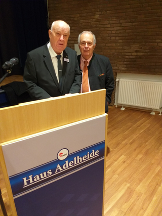 Zwei Männer stehen hinter einem 'Haus Adelheide' Rednerpult. Einer trägt einen Anzug, der andere eine Jacke und orange Krawatte.