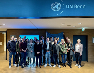 Eine vielfältige Gruppe Studierender und Besucher posiert am UN-Hauptsitz Bonn, mit UN-Logo und Flagge.
