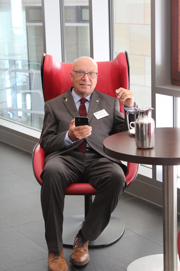 Ein lächelnder Mann im Anzug, Roland Heckenlauer, sitzt in einem roten Sessel und hält ein Smartphone.