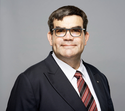 Kopfportrait eines Mannes mit dunklem Haar und Brille, der einen Nadelstreifenanzug und eine gestreifte Krawatte trägt und leicht lächelt, vor grauem Hintergrund.