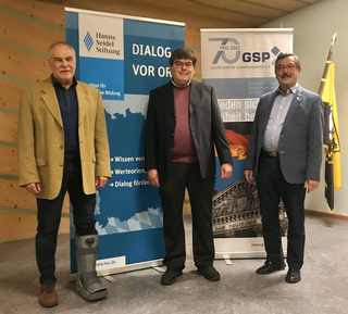 Drei Männer stehen vor Bannern der Hanns Seidel Stiftung und der Gesellschaft für Sicherheitspolitik; einer trägt einen orthopädischen Stiefel.