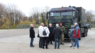 Ein Soldat in Tarnuniform erklärt einer Gruppe von Zivilisten einen großen Militär-LKW auf einem Außengelände.