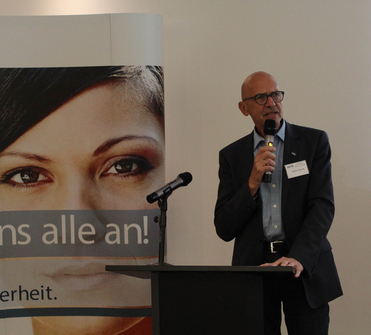 Reiner Schwalb spricht an einem Rednerpult in ein Mikrofon. Dahinter ein großes Banner mit Frauenporträt und Text.