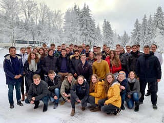 Gruppenfoto von Schülern und Erwachsenen lächelnd in einem verschneiten Wald, mit einem Bus in der Ferne.
