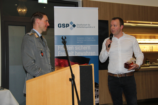 Ein Offizier und ein Redner mit Mikrofon und Geschenk vor einem GSP-Banner mit der deutschen Flagge.