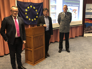 Drei Männer – einer im Anzug, einer zivil, einer in Militäruniform – stehen vor einer EU-Flagge und einer Präsentationsleinwand.