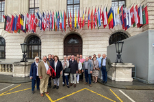 Eine Gruppe Erwachsener lächelt vor einem stattlichen Gebäude, das mit einer langen Reihe internationaler Flaggen geschmückt ist.