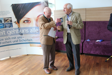 Zwei lächelnde ältere Männer, einer im Janker, übergibt dem anderen mit Mikrofon einen Gegenstand. Ein deutsches Banner im Hintergrund.