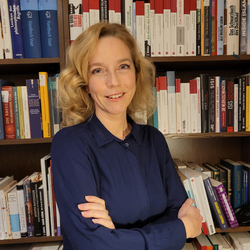 Eine lächelnde blonde Frau in einem dunkelblauen Hemd mit verschränkten Armen steht vor Bücherregalen voller Sachbücher.