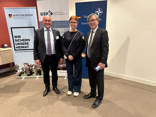 Drei Personen – zwei Männer, eine Frau – posieren vor Bannern des Reservistenverbands, der GSP und der Deutschen Gesellschaft.