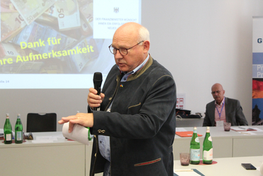 Ein kahler Mann in dunkler Jacke mit deutscher Flagge spricht in ein Mikrofon; im Hintergrund eine Präsentation mit Euro-Banknoten.