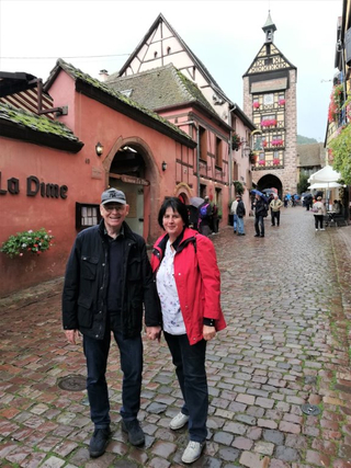 Ein Paar hält Händchen auf einer Kopfsteinpflasterstraße mit historischem Uhrenturm und Fachwerkhäusern im Hintergrund.