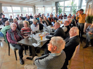 Ein Raum voller Gäste, hauptsächlich Senioren, sitzt an Tischen in einem hellen Restaurant oder Saal bei einem Vortrag.