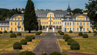 Ein prächtiger gelber Palast mit dunklem Dach und weißen Fenstern, umgeben von einem formalen Garten mit Zufahrt und geschnittenen Büschen.