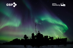 Leuchtende grüne und violette Nordlichter über einem dunklen Schiffswrack unter klarem Sternenhimmel.