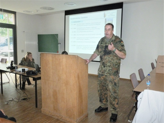 Militäroffizier präsentiert an einem Rednerpult in einem Klassenzimmer mit Projektionswand und einer Soldatin am Laptop.