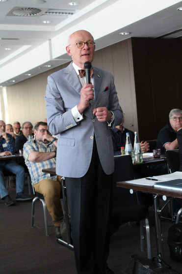 Ein kahler Mann in hellblauem Anzug mit Fliege spricht mit Mikrofon bei einer Konferenz vor einem sitzenden Publikum.