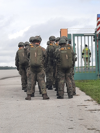 Soldaten mit Helmen und Rucksäcken stehen auf einem befestigten Platz, vor einem Tor mit sichtbarem Feuerwehrmann dahinter.