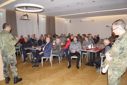 Ein Soldat in Tarnuniform spricht vor einem Publikum aus Militärangehörigen und Zivilisten an Tischen.