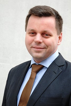 Professionelles Porträt eines lächelnden Mannes mit kurzen dunklen Haaren, im dunklen Anzug und brauner Krawatte.
