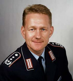 Lächelnder Mann in dunkler Uniform mit Schulterabzeichen vor schlichtem Hintergrund.