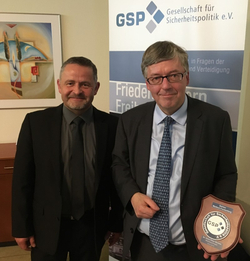 Zwei lächelnde Männer; einer hält eine GSP-Gedenktafel vor einem Banner der Gesellschaft für Sicherheitspolitik.