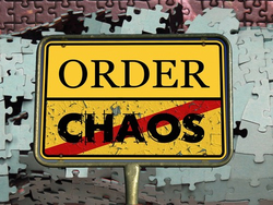 Ein gelbes Schild mit 'ORDER' oben und 'CHAOS' unten, rot durchgestrichen, vor einem Hintergrund aus Puzzleteilen.
