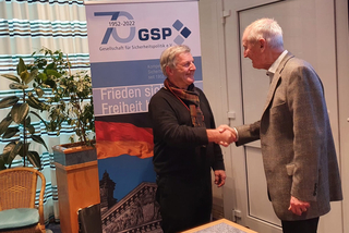 Zwei Männer schütteln sich die Hände vor einem Banner der Gesellschaft für Sicherheitspolitik (GSP) zum 70-jährigen Jubiläum.