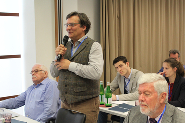 Ein Mann mit Brille und Weste spricht auf einer Konferenz in ein Mikrofon, im Hintergrund sitzen weitere Teilnehmer an Tischen.
