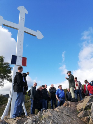 Gruppe lauscht Redner auf felsigem Hügel neben großem weißem Kreuz mit französischer Flagge bei blauem Himmel.