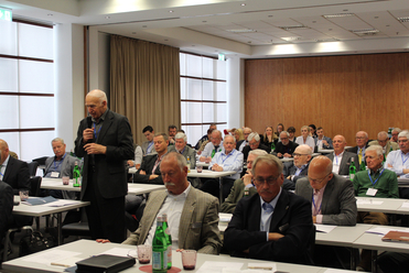 Ein älterer Mann spricht mit einem Mikrofon vor einem Publikum, das an Tischen in einem Konferenzraum sitzt.