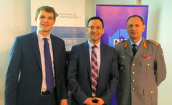 Drei Männer, zwei in Anzügen und einer in Militäruniform, lächeln vor Veranstaltungsbannern.