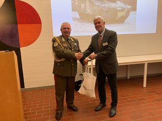 Zwei Männer, einer in Militäruniform und einer in Zivilkleidung, schütteln sich die Hände. Ein Panzer wird im Hintergrund projiziert.