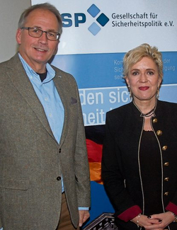 Zwei lächelnde Personen, ein Mann im Tweed-Sakko und eine Frau mit blonden Haaren, vor einem Sicherheitspolitik-Banner.