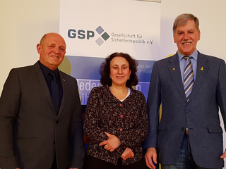 Drei lächelnde Personen, zwei Männer und eine Frau, vor einem Banner der GSP (Gesellschaft für Sicherheitspolitik e.V.).