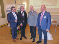 Vier lächelnde Männer, einer in Militäruniform, stehen bei einer GSP-Veranstaltung mit Bildschirm über Sicherheit und Verteidigung.