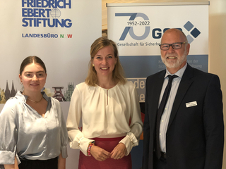 Drei Personen lächeln auf einer Veranstaltung vor Bannern der Friedrich-Ebert-Stiftung und der GSP zum 70. Jubiläum.