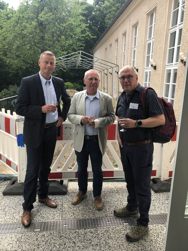 Drei Männer bei einer Veranstaltung im Freien, mit Getränken und Baustellenabsperrungen im Hintergrund.