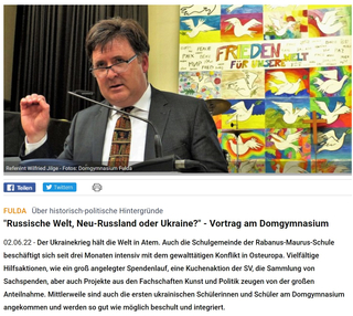Wilfried Jilge spricht vor einer bunten Wandcollage mit Friedenstauben und dem Schriftzug "FRIEDEN FÜR UNSERE WELT".