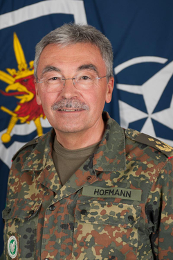Deutscher Militäroffizier HOFMANN in Flecktarn-Uniform, lächelnd vor Bundeswehr- und NATO-Flaggen.